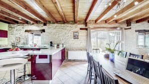 A rustic kitchen with stone walls, wooden beams, a large island with a sink, and a dining area with chairs and a laptop. - Gîte Ventoulou - Thegra (THEGRA)