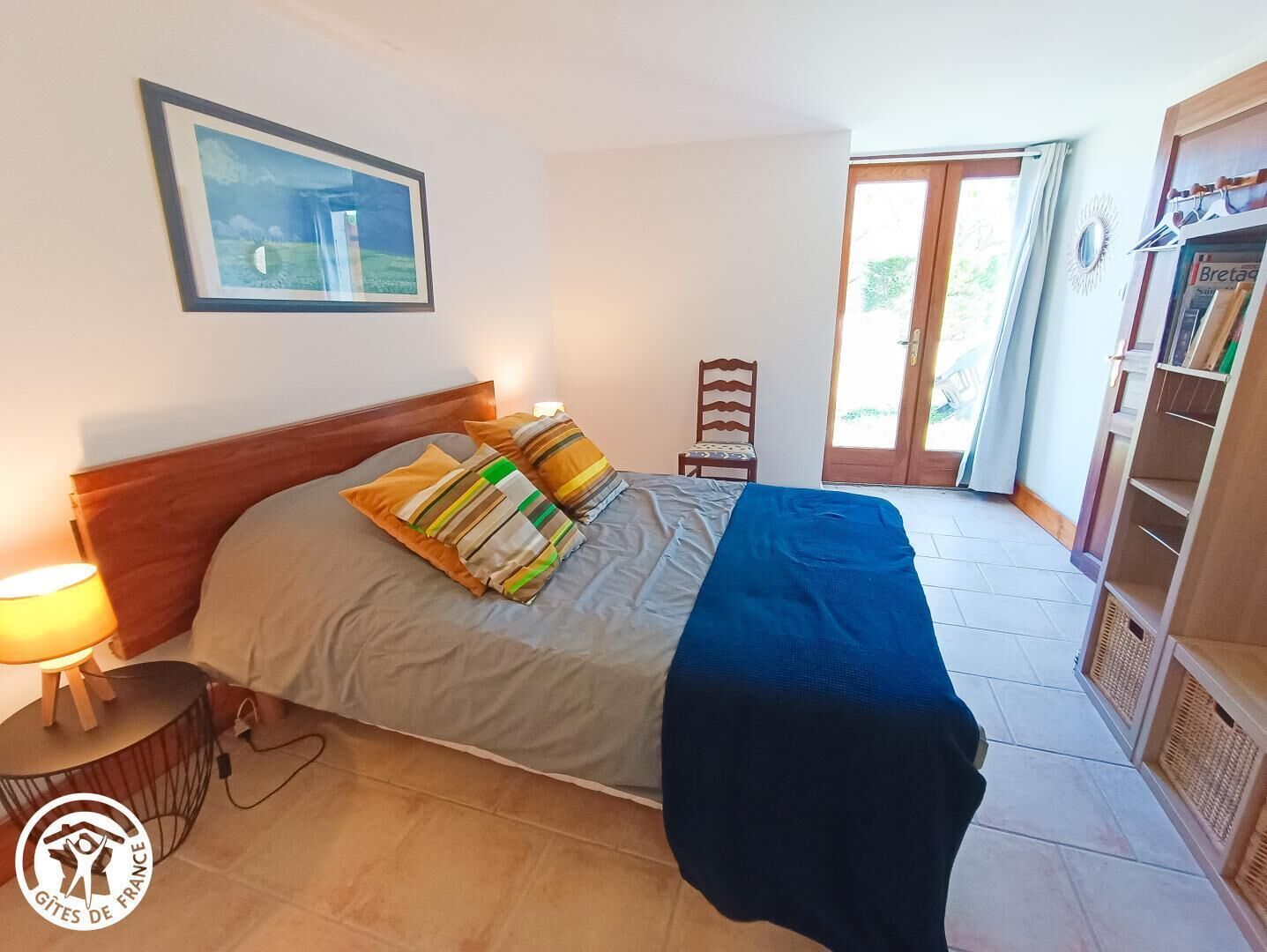 Un dormitorio con cama, mesita de noche, silla, puerta y estante con libros.