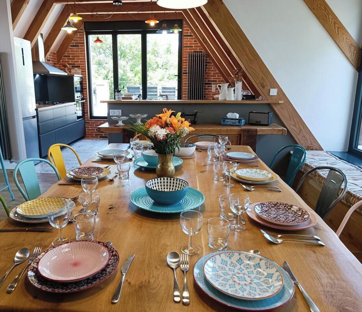 A dining area with a long table set for a meal, a kitchen in the background, and a view of the outdoors.