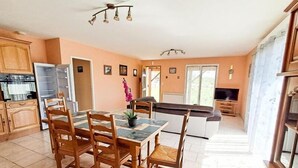 A spacious kitchen with wooden cabinets, a dining area with a tiled table and chairs, and a living area with a sofa and TV. - Gîtes de France® - Le TRUFFADOU (SAUZET)