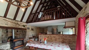 Un interior rústico con pared de piedra, vigas de madera, chimenea, una mesa de comedor con bancos y una cocina al fondo.