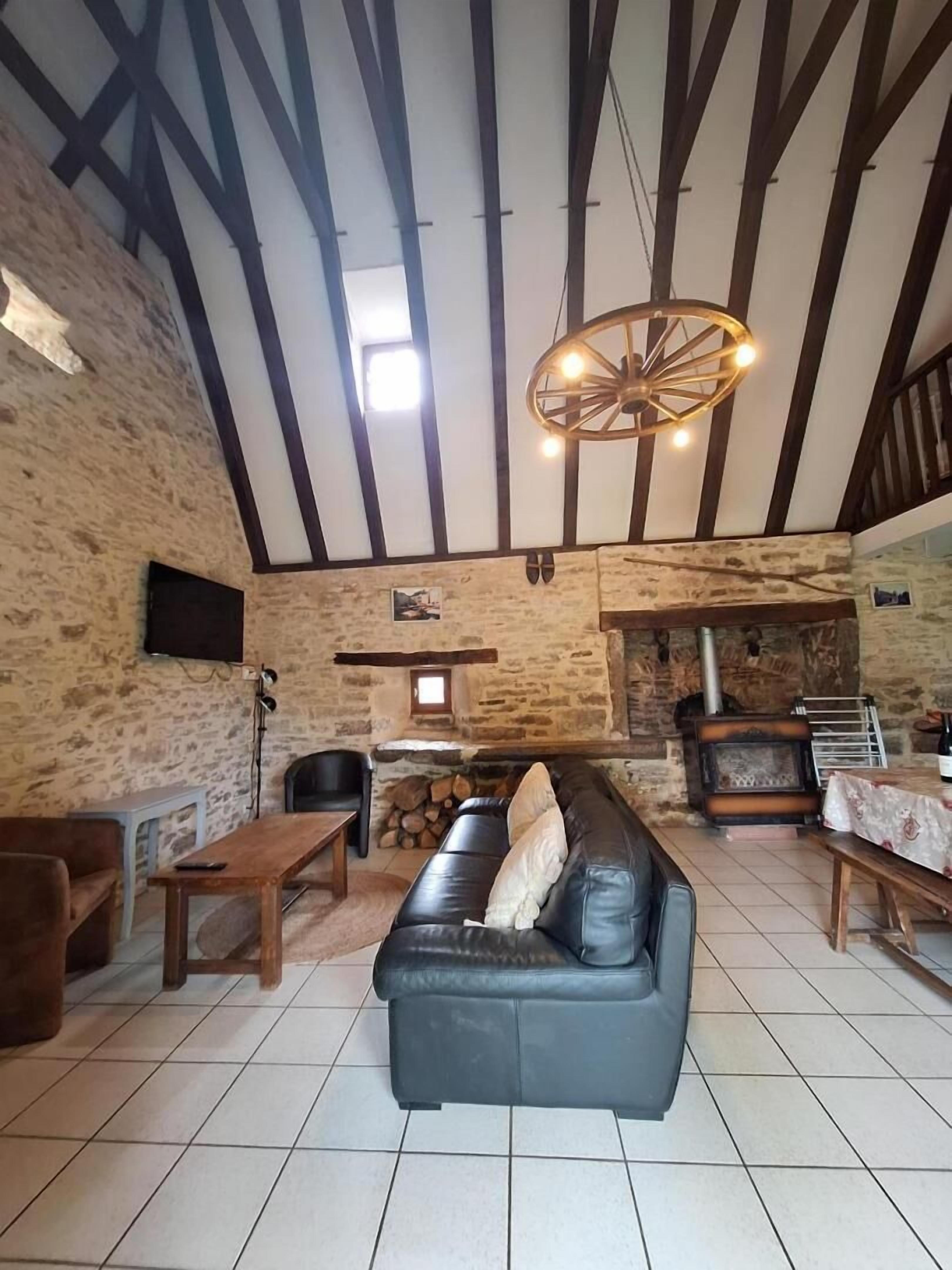 A rustic interior with exposed stone walls, a wooden ceiling, a black leather sofa, a wooden table, and a stone fireplace.