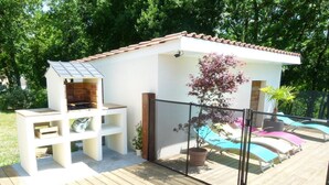 A wooden deck with a barbecue area, a glass-enclosed pool, and a small white building with a tiled roof. - Gîtes de France® - La Bouysse (SERIGNAC)
