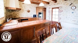 Une cuisine rustique avec des murs en pierre apparents, des poutres en bois, un îlot central recouvert de carreaux et un coin repas avec des chaises.