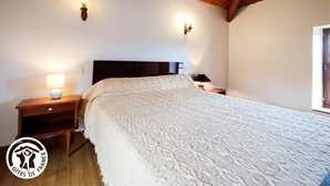 Une chambre avec un plafond en bois, un grand lit recouvert d’une couette blanche à texture, deux lampes fixées au mur et une table de chevet avec une lampe.