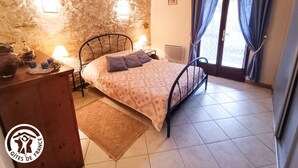 Une chambre avec un mur en pierre, un lit recouvert d’une couvre-lit à motifs, une commode en bois, une table de chevet avec une lampe, et une vue sur l’extérieur par une fenêtre ornée de rideaux.