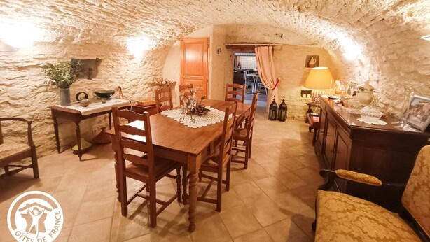 Un coin repas rustique avec une table et des chaises en bois, une petite cuisine à l’arrière-plan et un mur en pierre.