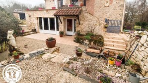 Une maison en pierre avec un toit en tuiles, un patio couvert et un jardin agrémenté de plantes en pot et d’une statue.