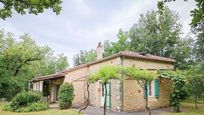 Una casa de piedra con techo de tejas, contraventanas verdes y un jardín con césped y árboles.