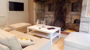 A modern living room with a stone fireplace, white furniture, and a flat-screen TV mounted on the wall. - Gîtes de France® - La Belle Lotoise (TEYSSIEU)