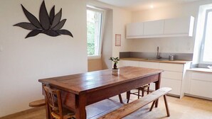 A dining area with a wooden table and benches, a small potted plant, and a kitchen in the background. - Gîtes de France® - La Belle Lotoise (TEYSSIEU)