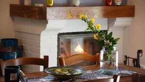 A dining table with a vase of yellow flowers, a patterned plate, and a candle holder. A fireplace with a visible fire is in the background. - Gîtes de France® - Gîte les Gratabels (BASTIT (LE))