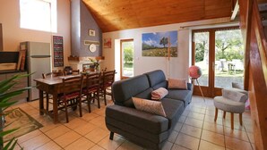 A cozy living room with a sofa, a dining table with chairs, a kitchen area, and large windows. - Gîtes de France® - Gîte les Gratabels (BASTIT (LE))