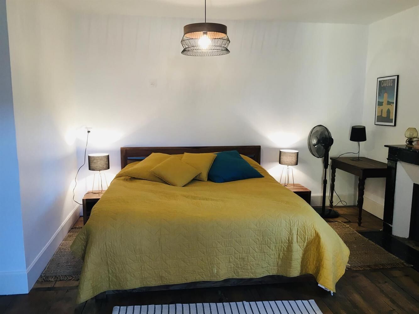 Une chambre avec un lit, deux lampes de chevet, un ventilateur sur pied et une petite table avec une lampe.
