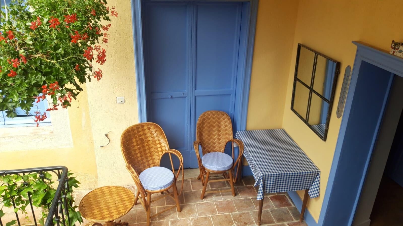 Ein kleiner Balkon mit zwei Korbstühlen und einem Tisch, eine blaue Tür und ein Fenster mit roten Blumen.