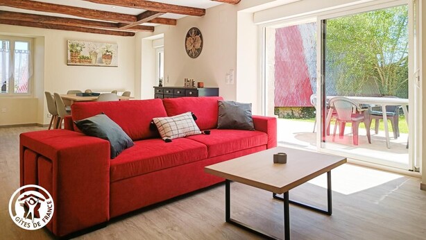 A spacious living room with a red sofa, a wooden coffee table, and a large sliding glass door leading to an outdoor area. - Gîtes de France® - Gîte du Fau (BESSONIES)