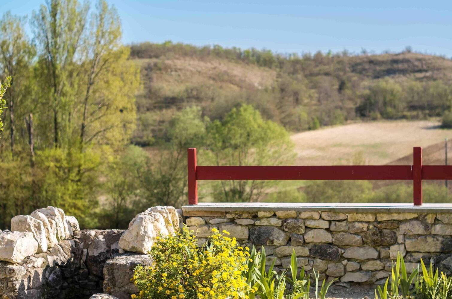 Un muro de piedra con cerca de madera roja, un sendero de piedra y vista a un paisaje montañoso con árboles.