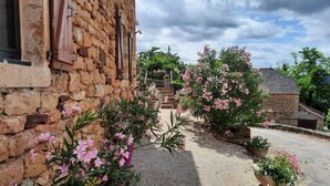 Un edificio rustico in pietra con persiane in legno, un vialetto ghiaioso e piante fiorite.