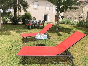 Ein Garten mit zwei roten Liegestühlen, einem Tisch mit Tablett und einem Gebäude im Hintergrund.