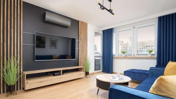 A modern living room with a flat-screen TV, a blue sofa, a round coffee table, and a large window with curtains.
