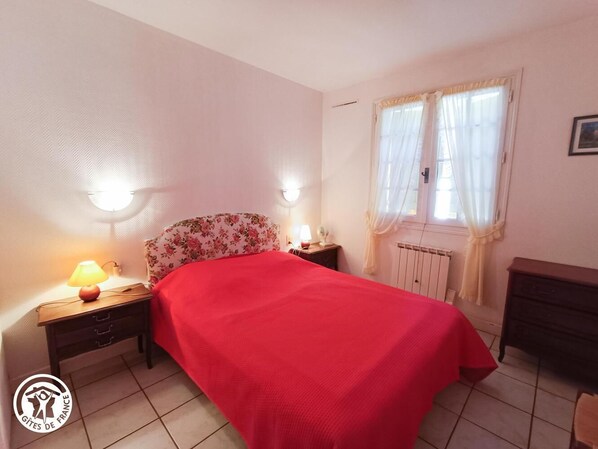 Une chambre à coucher avec une couvre-lit rouge, deux lampes de chevet, une tête de lit à motifs floraux, un radiateur et une fenêtre avec des rideaux légers.