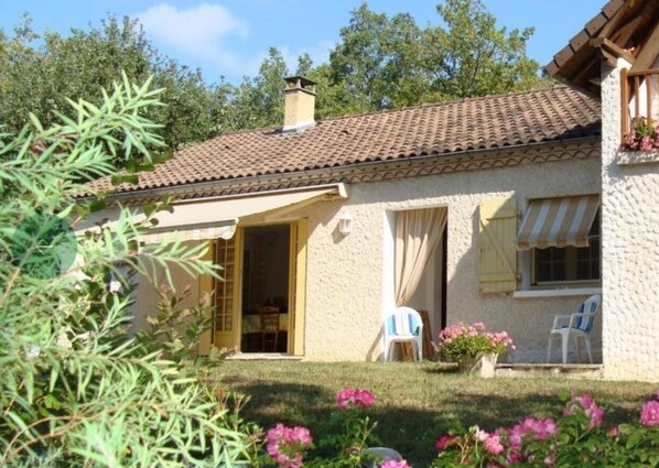 Ein Haus mit Ziegeldach, einem zur überdachten Terrasse führenden Eingang und einem kleinen Garten mit rosa Blüten.