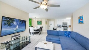 A modern living room with a large flat-screen TV, a blue sofa, a ceiling fan, and a kitchen area in the background. - Best view in Aventura Yacht Club, 2-bedroom condo fully equipped  (Aventura)