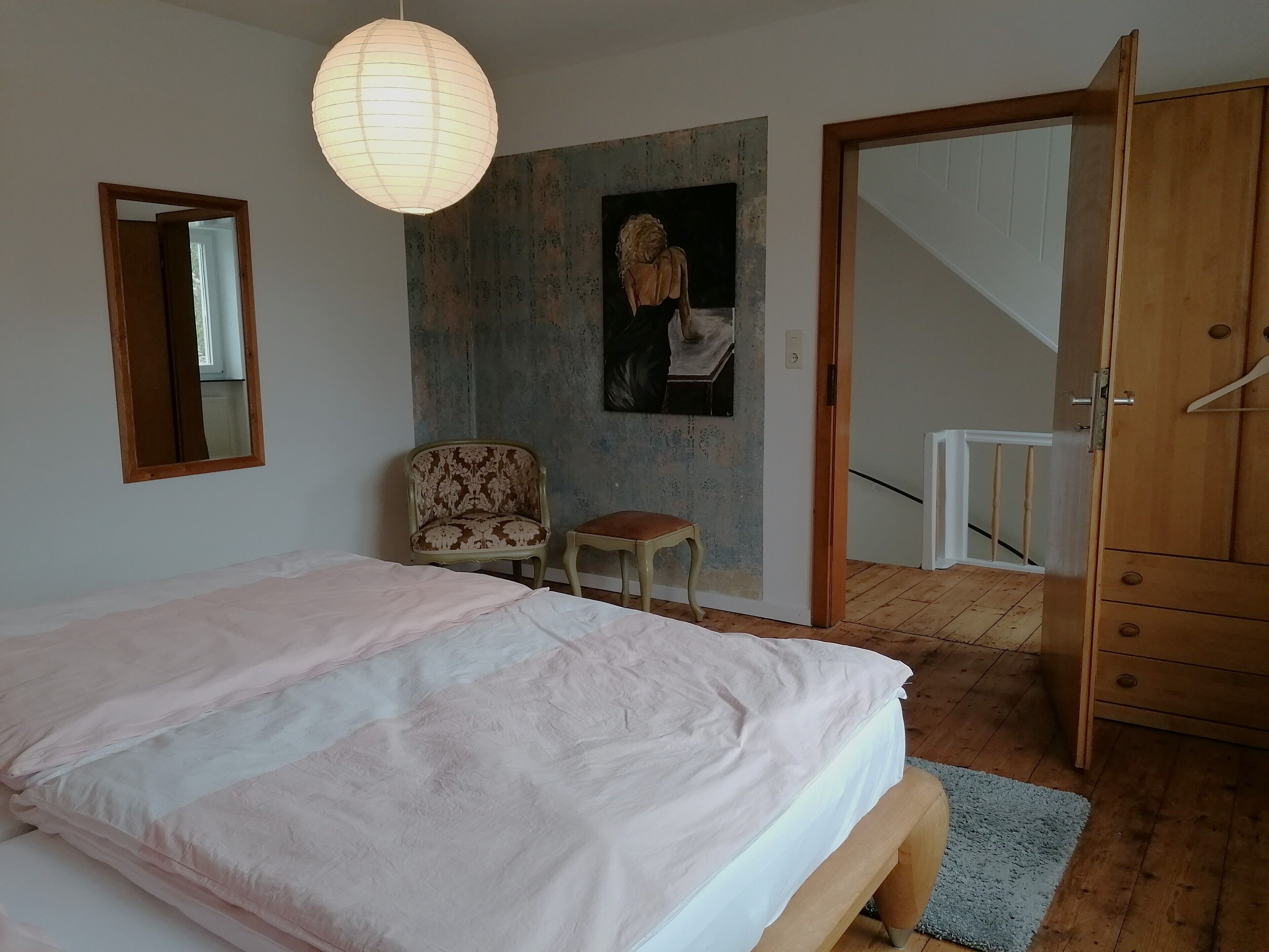 A bedroom with a bed, a chair, a wooden wardrobe, a mirror, and a wall with a painting.