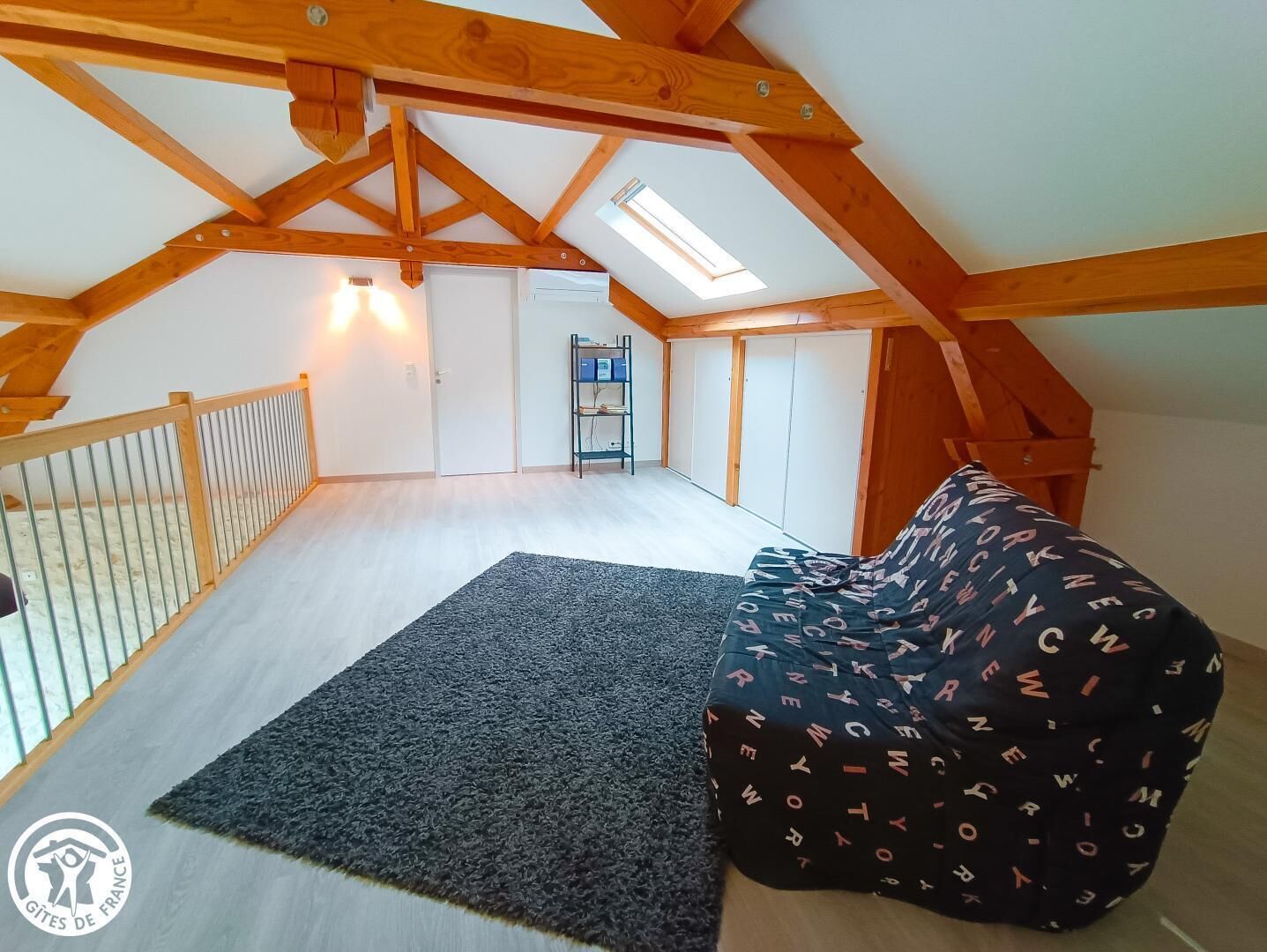 A spacious attic room with a high ceiling, wooden beams, a skylight, a black sofa, a small rug, and a metal railing.
