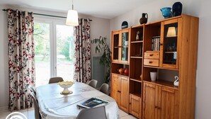 A dining area with a table set for two, a wooden cabinet with glass doors, and a view of the outdoors through a window.