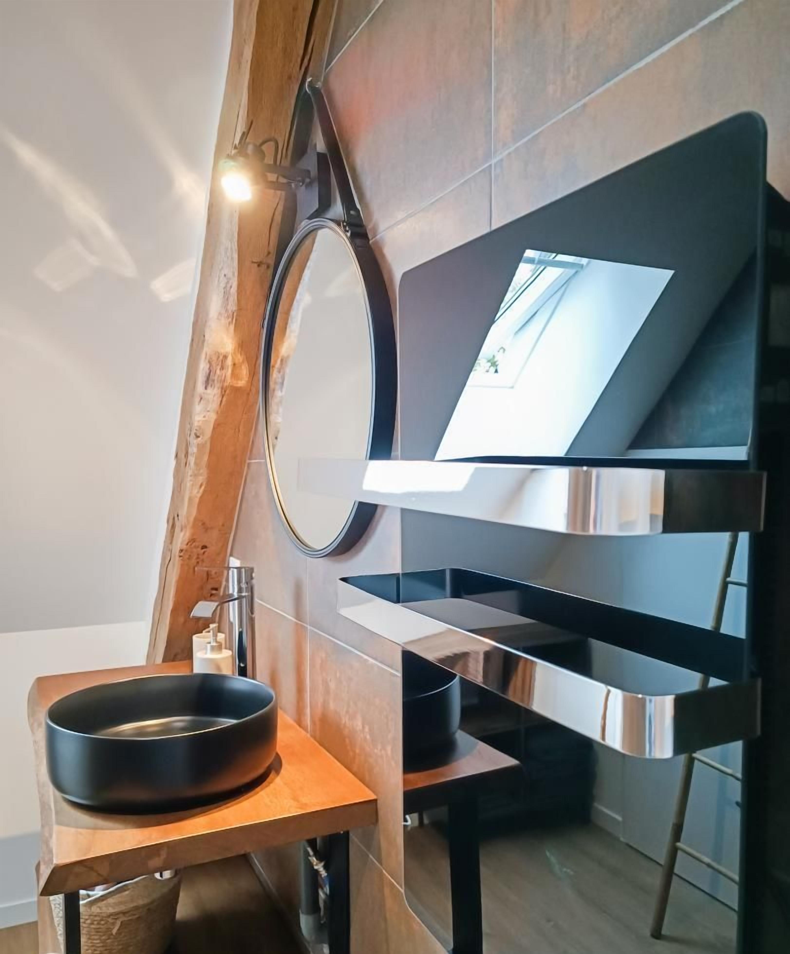 A modern bathroom with a wooden sloped ceiling, a round mirror, a black basin, and a built-in shelf.