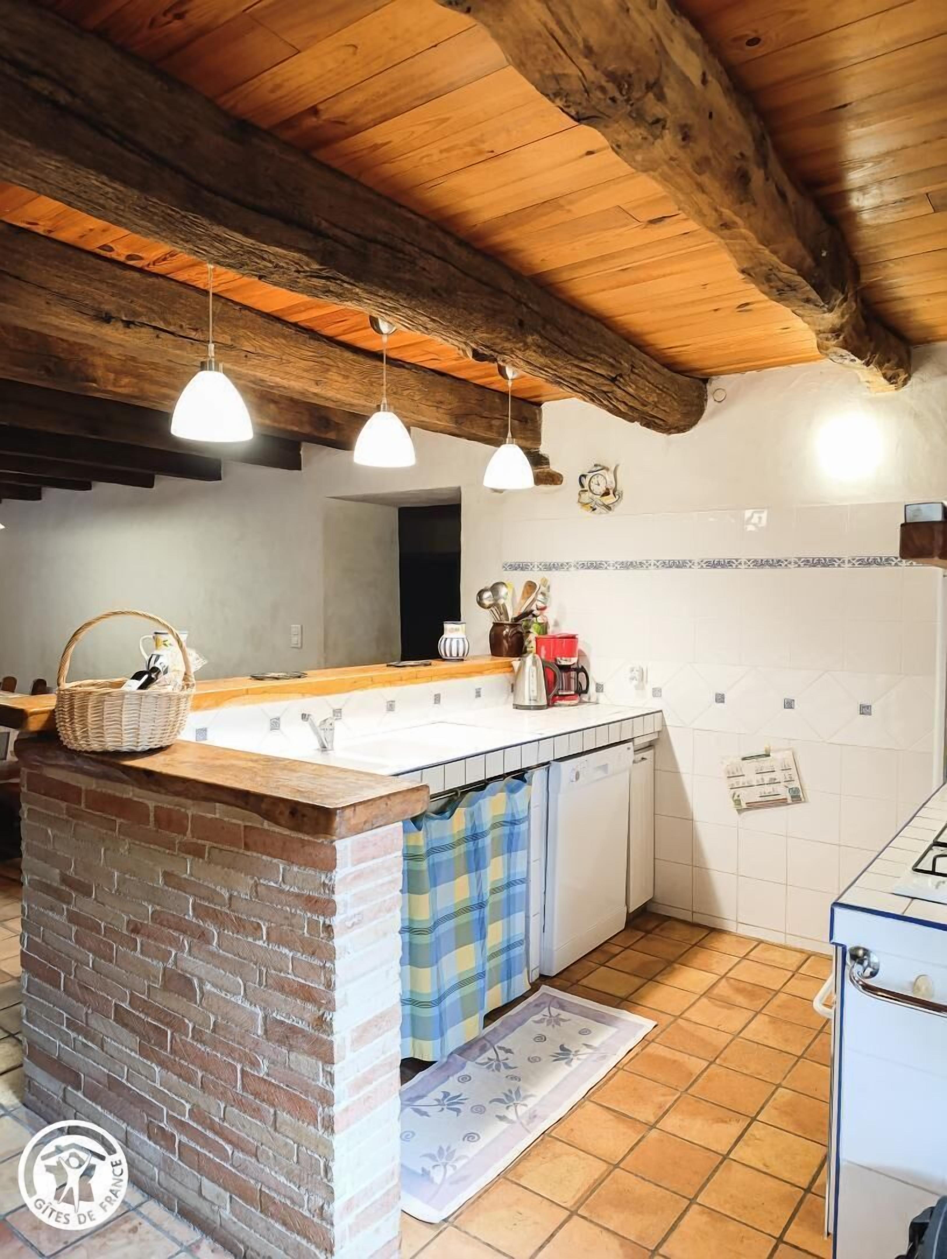Une cuisine dotée d’un îlot central en briques, de poutres apparentes en bois au plafond, de luminaires suspendus et d’un mur recouvert de carreaux.