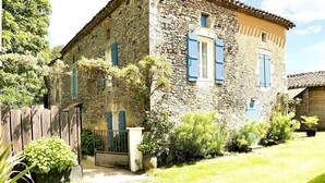 Un bâtiment en pierre, avec une clôture en bois, un portail et un petit jardin où poussent différentes espèces de plantes.