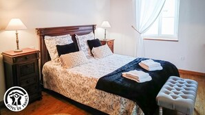 Une chambre à coucher avec une tête de lit en bois, deux tables de chevet avec des lampes, un grand lit avec une literie à motifs floraux, un banc au pied du lit et une fenêtre avec des rideaux légers.
