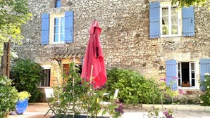 Un bâtiment en pierre avec des volets bleus, un parasol rouge et des plantes en pot.