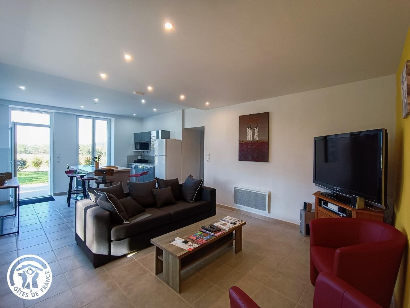 Un salon moderne avec un canapé, une table basse, un téléviseur et un fauteuil rouge. On aperçoit une cuisine à l’arrière-plan, avec un coin repas.