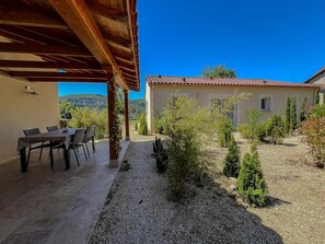 A covered patio with a table and chairs, a gravel garden with shrubs, and a building with a tiled roof. - Gîtes de France® - La Vigne Blanche (TOUR DE FAURE)