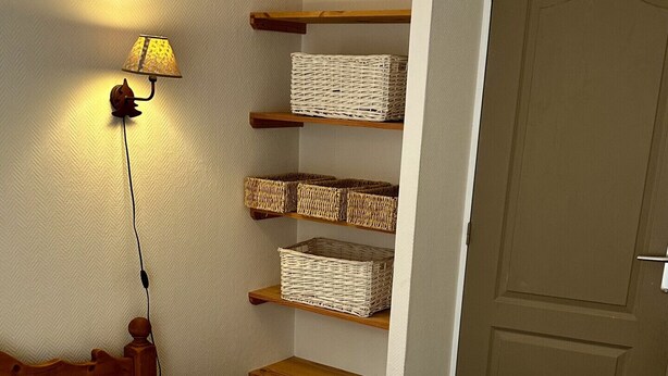 Ein ordentlich bezogenes Bett mit weißen Leinen und Kissen, Holzregale mit Körben, eine Wandlampe und eine geschlossene Tür.