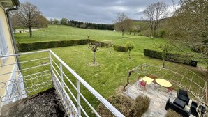 Ein Balkon mit Blick auf eine grüne Rasenfläche, eine Steinterrasse mit Gartenmöbeln und ein Heckenstreifen.