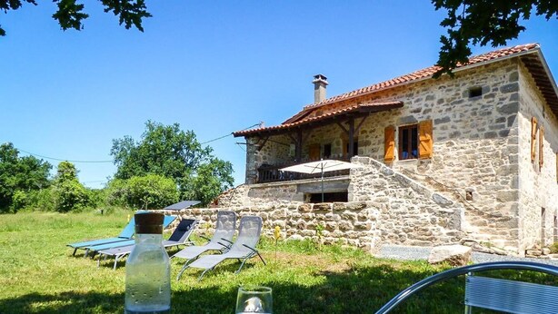 Une maison en pierre avec un toit en tuiles, un petit espace extérieur aménagé avec des chaises et une table, et une bouteille d’eau posée sur la table.