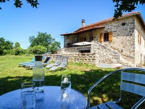 A stone house with a tiled roof, a small outdoor seating area with chairs and a table, and a bottle of water on the table. - Gîtes de France® - L'Oustalou (SAINT-BRESSOU)