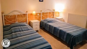Tweepersoonskamer met twee bedden, elk voorzien van een gestreept laken, een nachtkastje met lamp en een klein ladeblok. De kamer heeft een houten plafond en een radiator aan de muur.