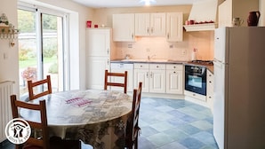 Een keuken met witte kastjes, een eettafel met stoelen, een koelkast en een raam met uitzicht op groen.