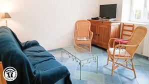 Een woonkamer met een bank, twee rieten fauteuils, een glazen salontafel, een houten kast met een televisie en een klok aan de muur.