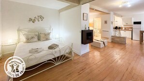 Een ruime slaapkamer met een groot bed, een klein nachtkastje en een ingebouwde televisie. Er is een keukenhoek met witte kasten en een koelkast.