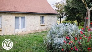 Ein Steinhaus mit rotem Dach, zwei Fenster mit Fensterläden und ein Garten mit Sträuchern und roten Blüten.
