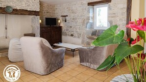 Ein Zimmer mit Steinwänden, einem Fernseher auf einem Holzkasten, zwei grauen Sesseln, einem kleinen Tisch und einer großen Pflanze.
