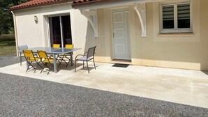 Uma casa térrea com telhado de cerâmica, um alpendre com mesa e cadeiras, e uma porta que dá para o interior.