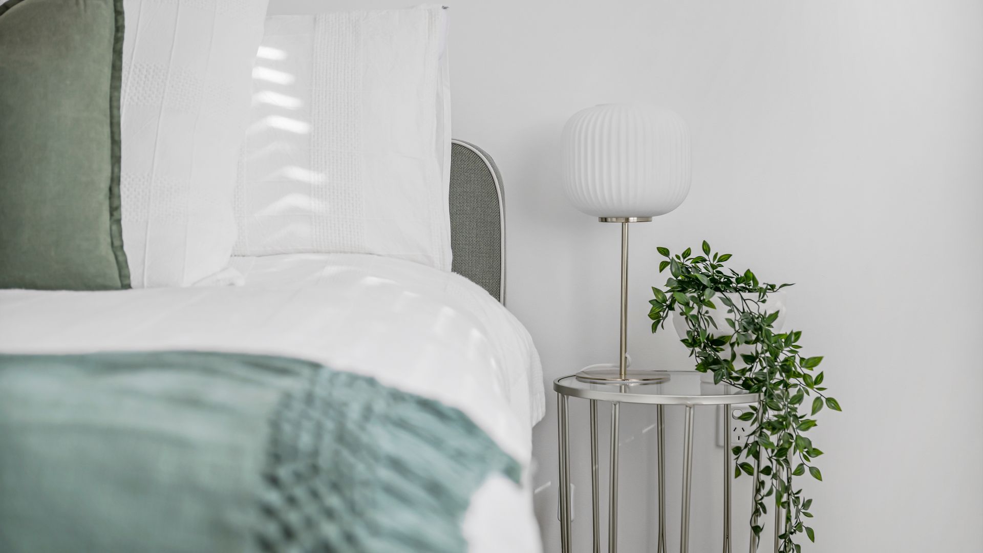 A neatly made bed with a green throw, a bedside lamp, and a potted plant on a bedside table.