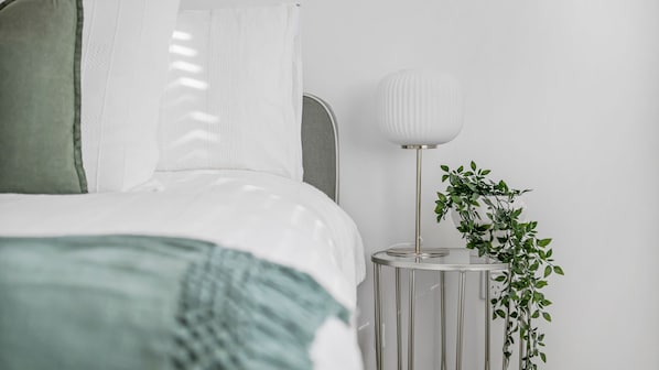 Una cama bien hecha con una colcha verde, una lámpara de noche y una planta en maceta sobre una mesita.